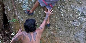 A father is keeping a close watch over his climbing son on an outdoor boulder. He stands braced, ready to assist his child if needed.