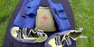 Picture of two knee support bandages with a pair of aggressive climbing shoes and a personal first aid kit spread out on a bouldering pad.