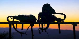 Picture of two backpacks balancing on a railing in the foreground with a orange sunset sky illuminating the hills in the distance.